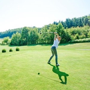 A female golfer performs a powerful tee-off on the expansive green fairway of the Stegersbach golf course in front of a forest backdrop.