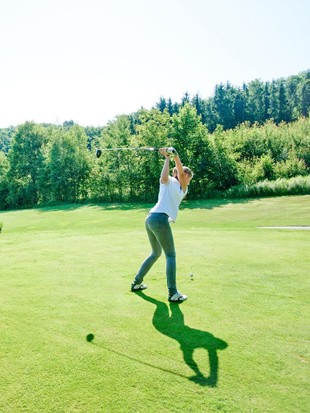 A female golfer performs a powerful tee-off on the expansive green fairway of the Stegersbach golf course in front of a forest backdrop.