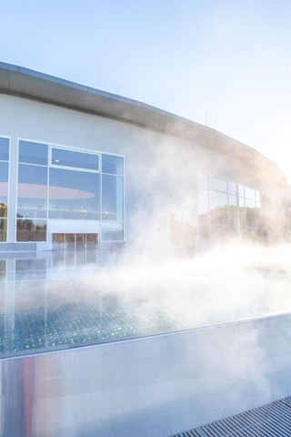 Enjoy soothing relaxation in the warm thermal water A modern outdoor pool with steaming thermal water in front of a large glass facade, reflecting the warm sunlight.