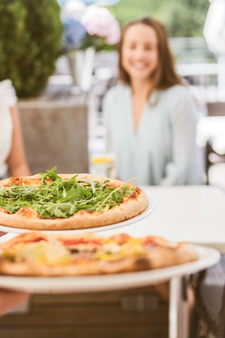 Enjoy a taste of Italy A waiter serves two freshly baked Sicilian pizzas topped with arugula on a sunny outdoor terrace, while two guests wait smiling in the background.