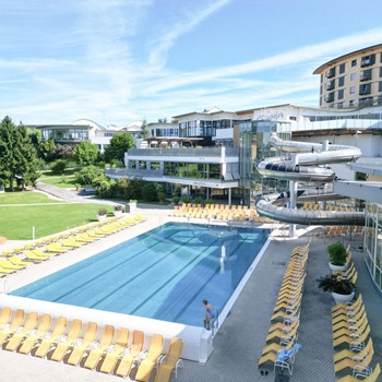 Weitläufige Außenanlage der Therme Stegersbach mit einem großen Sportbecken, zahlreichen gelben Liegestühlen, einer spiralförmigen Röhrenrutsche und dem Hotelgebäude im Hintergrund.