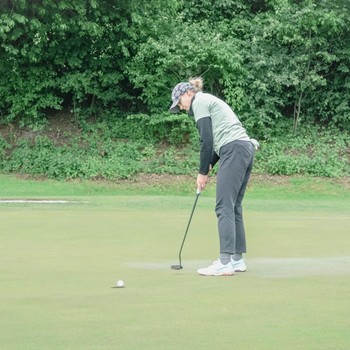 A female golfer putting on the green, aiming to sink the ball into the hole, surrounded by trees and lush landscape.