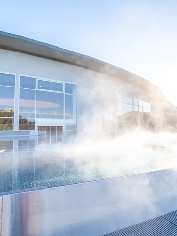 Ein moderner Außenpool mit dampfendem Thermalwasser vor einer großen Glasfassade, in der sich das warme Sonnenlicht bricht.