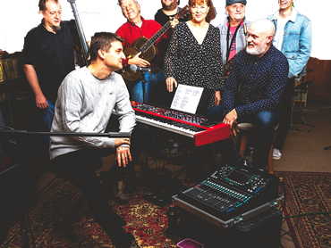 Eine achtköpfige Band mit Sängern und Musikern an Keyboard, Gitarren und Saxophon probt gemeinsam in einem gemütlichen Studio.