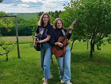 Zwei junge Frauen stehen lachend mit ihren Akustikgitarren auf einer grünen Wiese vor einer weiten Hügellandschaft.