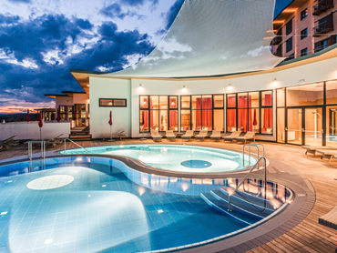 Die beleuchtete Außenpool-Anlage des Allegria Resorts Stegersbach mit Blick auf das Hotelgebäude während der Abenddämmerung.