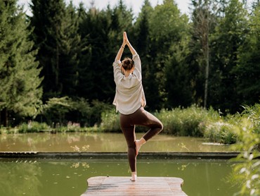 Eine Frau steht am Steg eines Teiches und macht eine Yogapose.