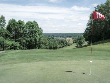 A golf course with a red flag on the 18th hole, surrounded by trees and gentle hills.