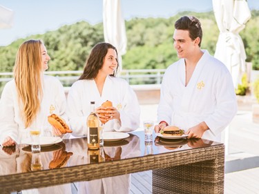 Drei junge Gäste in weißen Bademänteln genießen gemeinsam Burger und Hot Dogs an einem Tisch auf der sonnigen Außenterrasse des Thermen-Bistros.