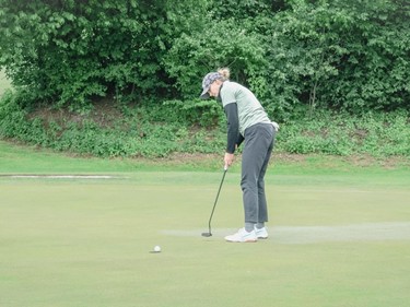 A female golfer putting on the green, aiming to sink the ball into the hole, surrounded by trees and lush landscape.