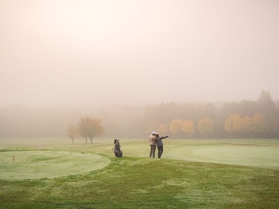 Ein Paar steht an einem nebligen Morgen auf dem grünen Rasen eines Golfplatzes und blickt gemeinsam in die weite, herbstliche Landschaft.