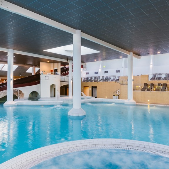 Swimming fun and relaxation in the extensive indoor thermal baths Spacious indoor thermal pools featuring bright columns, an integrated waterfall, and an adjacent water slide.