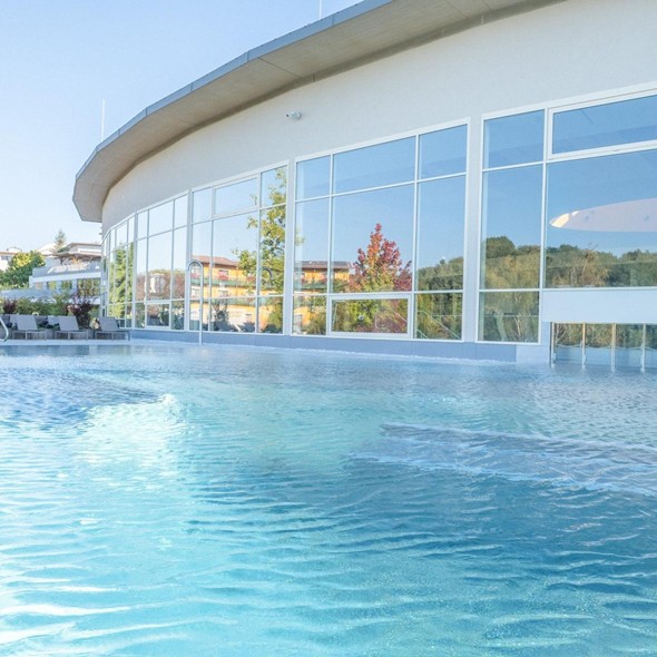 Large, light blue outdoor pool in front of a modern building with a curved glass façade on a sunny day.