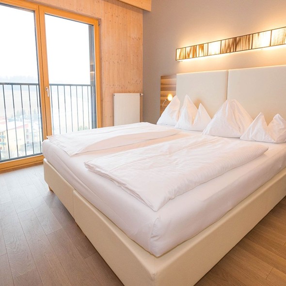 Natürliche Geborgenheit und helles Design erleben Modernes, helles Doppelzimmer aus heimischem Fichtenholz mit einem beigen Polsterbett, hellem Parkettboden und einer großen Fensterfront für viel natürliches Licht.